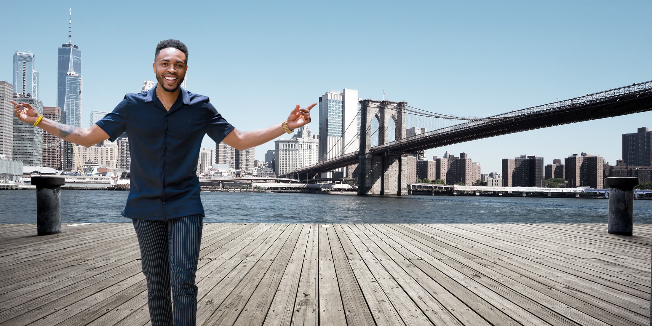 Antonio Edwards with arms outstretched on a Brooklyn pier, the Brooklyn Bridge and Manhattan skyline behind him
